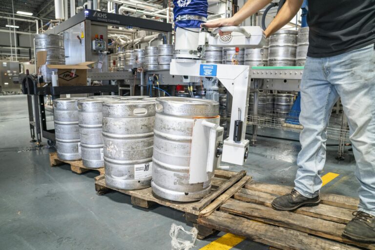A worker operates machinery to stack steel kegs in a brewery warehouse.