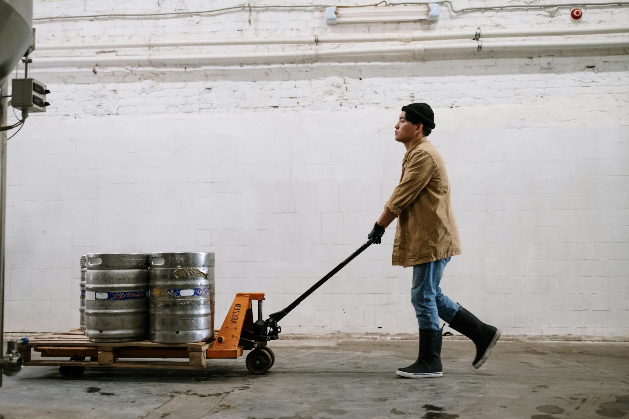 Brewery worker wearing gloves and boots moving metal kegs on a trolley in an industrial indoor space.